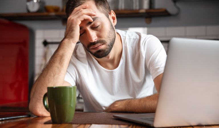 Man sitting in front of his laptop, wearing white t-shirt, having his hand on his forehead, feeling tired. Read the article: Will CBD Make You Tired?